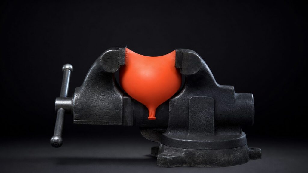 An orange balloon held in the jaws of a black bench vise.
