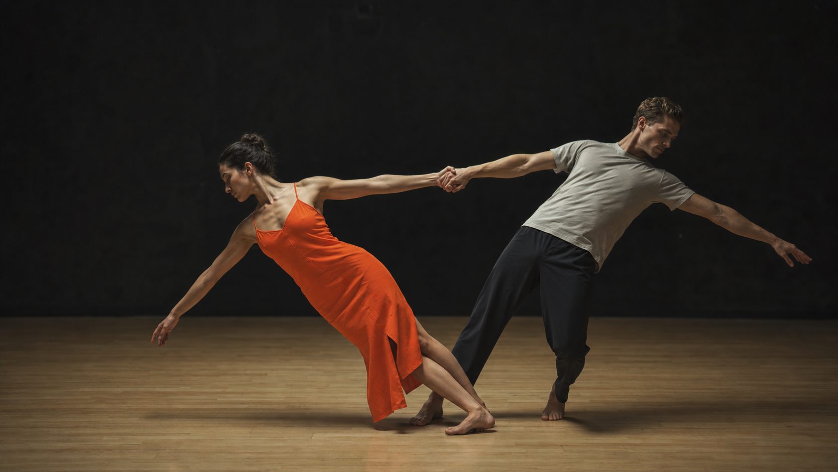 Two dancers in a counterbalance lean, one in an orange garment, on a bare stage.